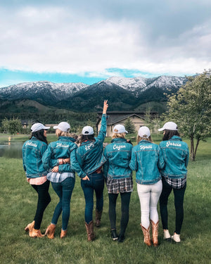 A group of women stand in front of the mountains wearing jean jackets customized with their names across their shoulders in white font.