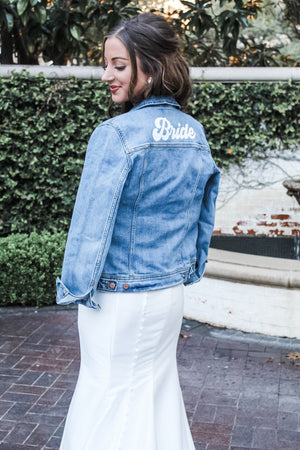 A bride wears a customized denim jacket which reads "Bride" in white text over her gown.