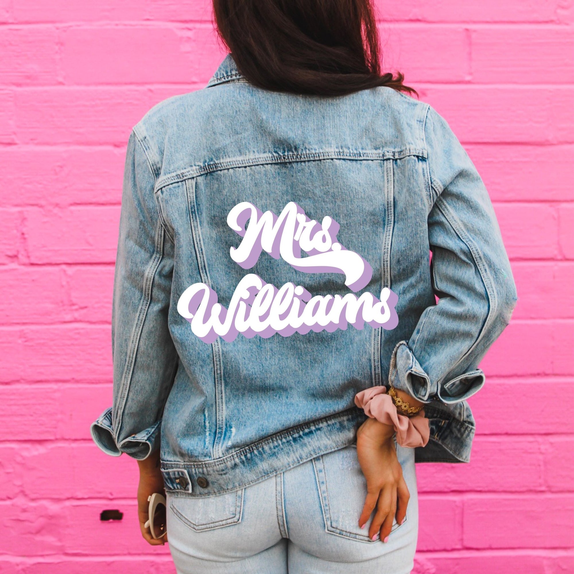 A woman stands in front in a pink wall showing off her customized denim jacket with her last name printed on it in pink and white.