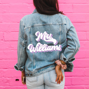 A woman stands in front in a pink wall showing off her customized denim jacket with her last name printed on it in purple and white.