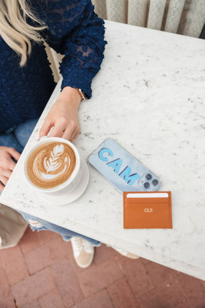 A monogrammed, tan cardholder and phone case sit on a table next to a latte
