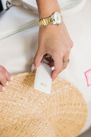 A person attaches a straw beach hat to their bag using a white hat clip with a gold foil monogram.