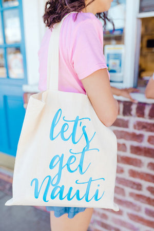 A woman stands holding a tote that reads "let's get nauti" in bright blue