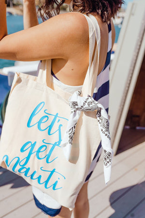 A woman stands holding a tote that reads "let's get nauti" in bright blue