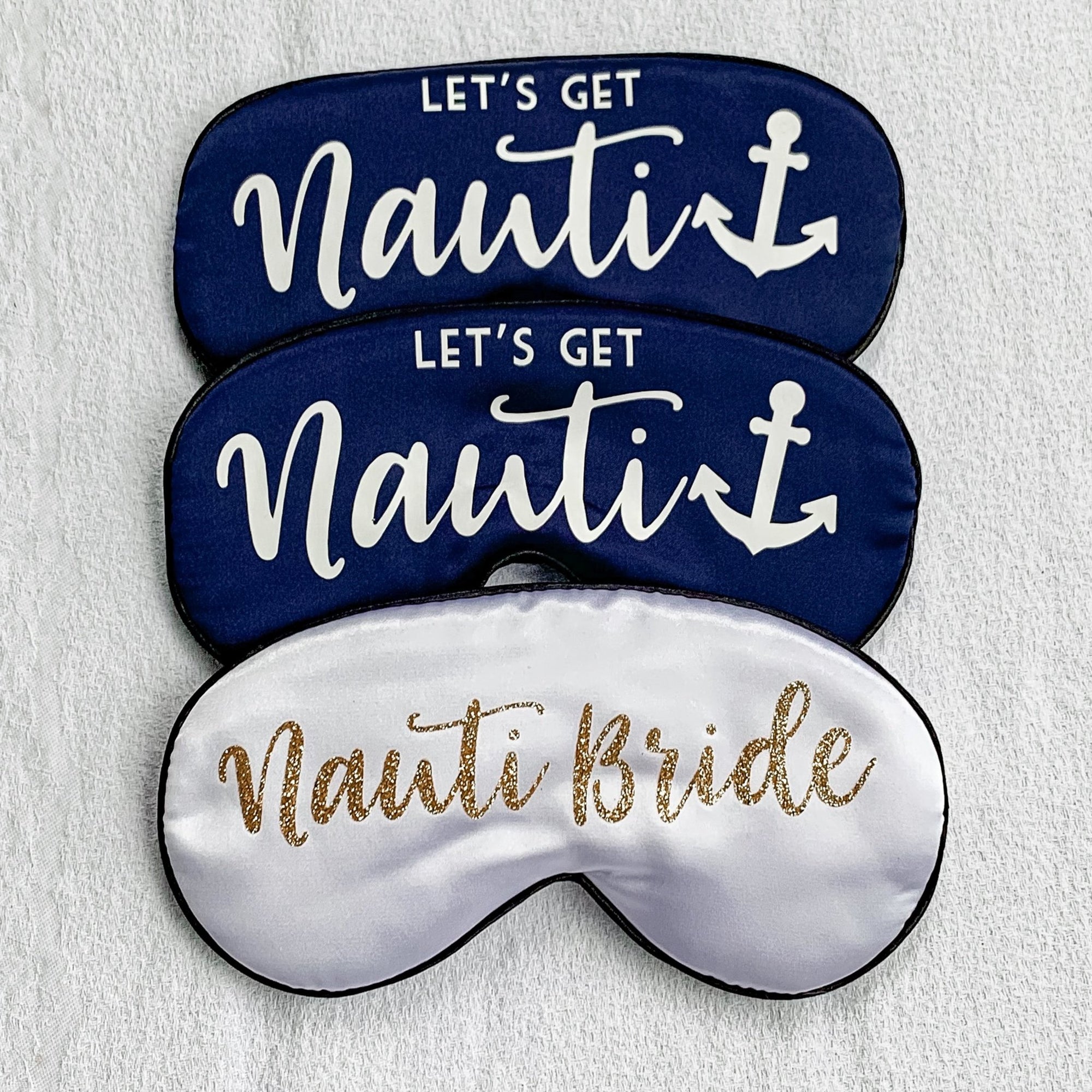 A white sleep mask that reads "Nauti Bride" in gold is paired with two navy sleep masks which read "Let's Get Nauti" in white writting.