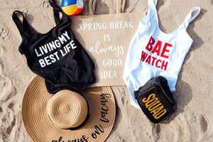 An assortment of products including a tote bag, swimsuits, a fanny pack, and a floppy beach hat sit on the sand at the beach.