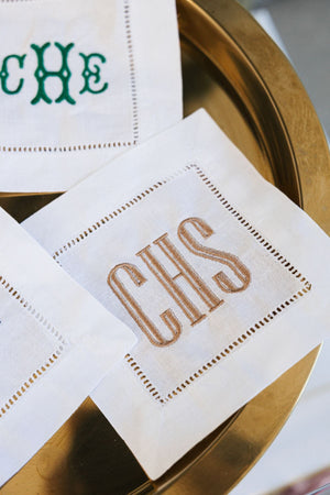 A cocktail napkin is shown up close to show off it's embroidered monogram in a beige thread color.