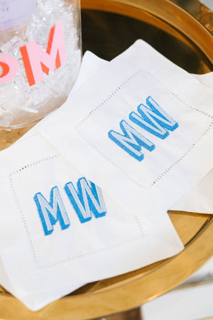 A set of cocktail napkins sits out showing off it's vibrant blue monogram which is embroidered onto the napkin.