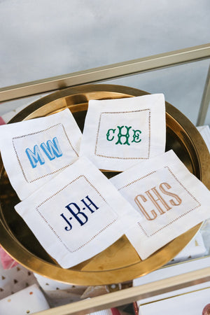 Four cocktail napkins are laid out on a bar cart to show some of the embroidered monograms that can be used to customize this product.