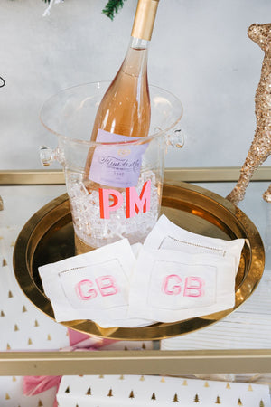 A set of cocktail napkins sits on a bar cart next to a wine bottle in an ice bucket with an embroidered pink monogram.