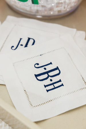 A set of cocktail napkins is embroidered with a monogram in a navy thread.