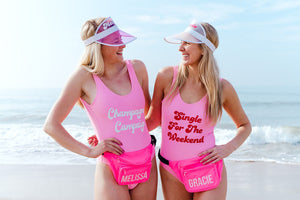 Two girls at the beach stand in their custom pink swimsuits and fanny packs which have their names printed on them.