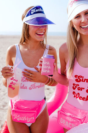 Two girls stand at the beach laughing while sporting their custom swimsuits, visors, and fanny packs.
