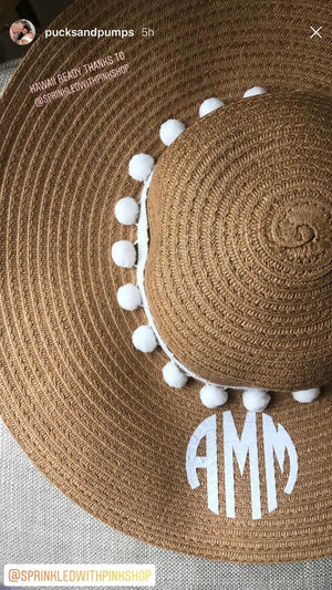 A floppy hat with white pom poms is customized with a white circular monogram.