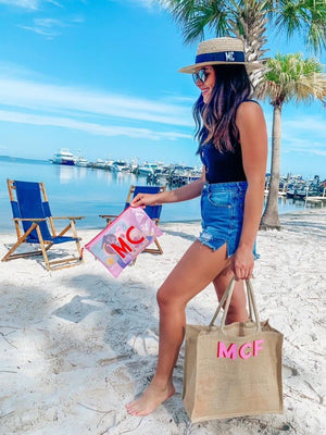 A woman walking on the beach holds a monogrammed pool bag and tote bag while wearing a monogrammed boater hat.