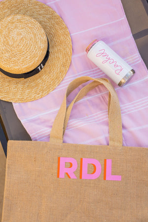 A personalized tote is laid out on a pink towel with a straw hat and custom drink cooler.