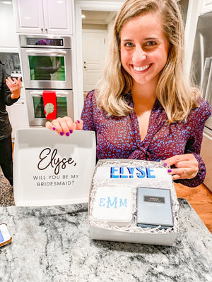 A woman holding a box that reads "Will you be my bridesmaid?" and contains our monogrammed jewelry case and customizable tumbler
