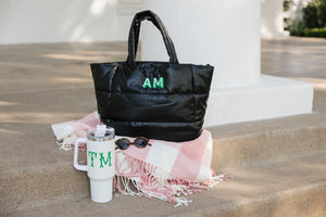 A black puffer tote sites next to a white monogrammed tumbler