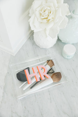 An acrylic catchall box is used for holding makeup brushes and is monogrammed with a pink and melon monogram.