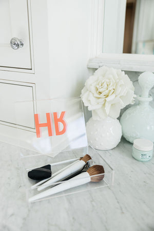 An acrylic catchall box is used for holding makeup brushes and is monogrammed with a pink and melon monogram.