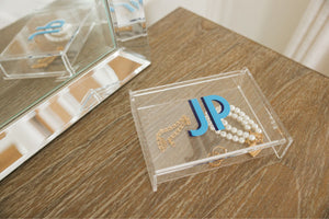 An acrylic catchall box is personalized with a blue and navy monogram and is used to collect rings and bracelets.