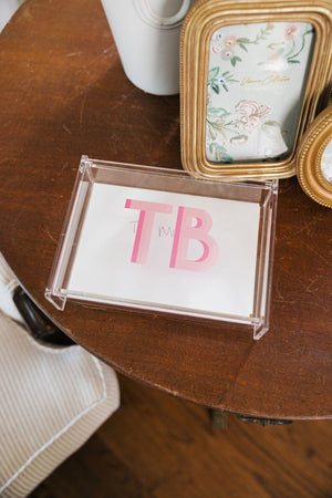 A pink monogrammed acrylic catchall box is used to collect cards on a brown wooden table.