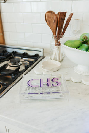 An acrylic catchall box is used to collect recipe cards in a kitchen and is monogrammed with purple letters.