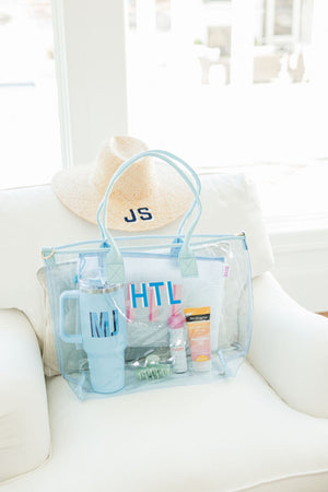 A blue clear tote bag is filled with beach essentials including a blue 40 oz tumbler and sunscreen.