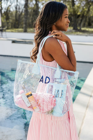 A woman in a pink striped dress holds a blue clear tote with a towel and a tumbler inside.
