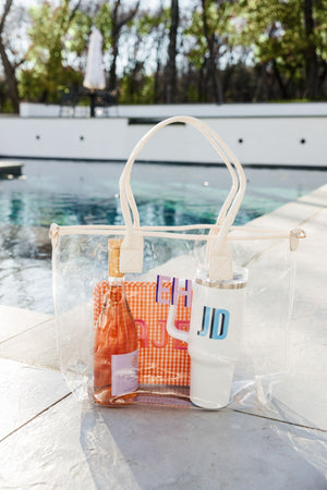 A clear tote bag with white trim is placed in front of the pool filled with a bottle of rose, an orange pouch, and a white 40 oz tumbler.