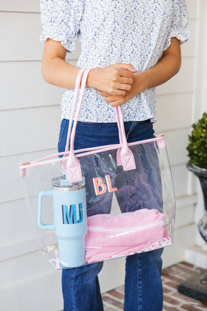 A person holds a pink clear tote bag with a blue 40 oz tumbler and a pink towel inside.