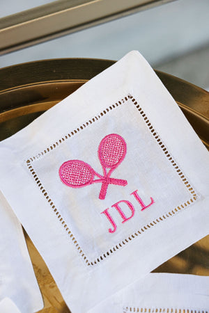 A cocktail napkin with two tennis rackets and custom monogram in pink thread.