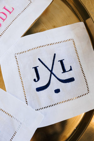 An embroidered cocktail napkin with hockey sticks and a custom monogram in navy thread.