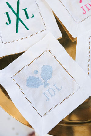A cocktail napkin with pickleball paddles and a custom monogram in light blue thread.