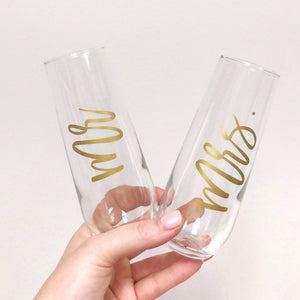 A person holds up two champagne flutes customized with "Mr." and "Mrs." in gold script font.