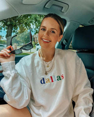 A young blonde sitting in a car wears a sweatshirt with "dallas" embroidered in multiple colors