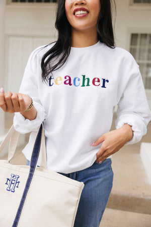 A woman wears an embroidered sweatshirt that reads "teacher" and holds a monogrammed canvas tote