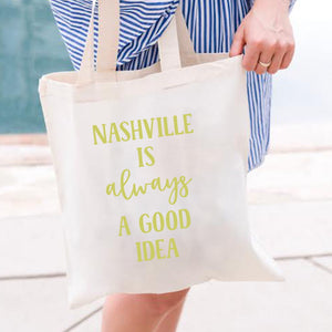 A woman holds a tote which reads "Nashville is always a good idea" in gold font.