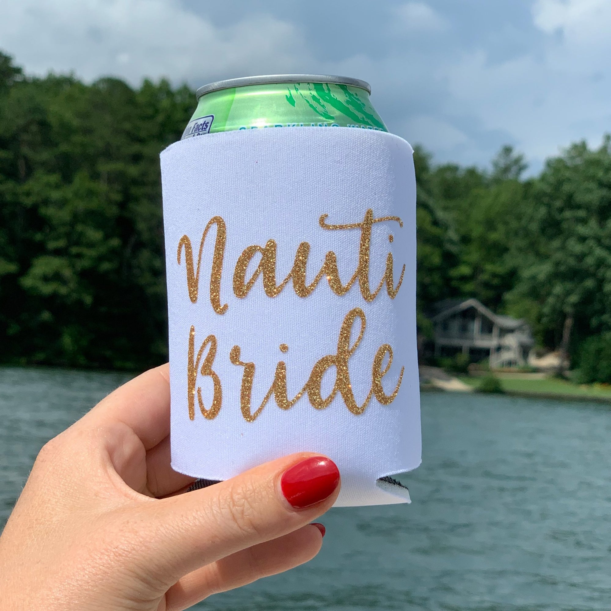 A woman holds up a white can cooler which says "Nauti Bride" in a gold glitter font.