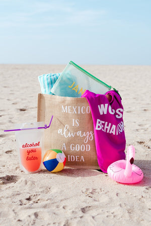 An assortment of products sits on the beach in or next to a tote which reads "Mexico is always a good idea."