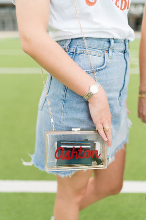 A girl on a field wears a clear stadium bag customized with her name in a red font.