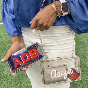 A woman wears a clear stadium bag customized with her name in a white font and holds a phone that has a monogrammed case.