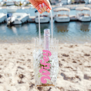 A person holds a wine bag on the beach which says "Wifey" in a pink font.