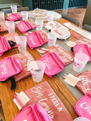 A group of mauve shirts and pink fanny packs are customized and laid out on a table for a bachelorette.