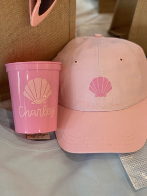 A pink baseball hat and a pink cup are customized with a pink seashell design