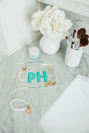 A mint and turquoise monogrammed acrylic tray is placed in a bathroom with some jewelry.