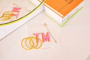 An acrylic tray is customized with a pink monogram and is used to hold gold jewelry.