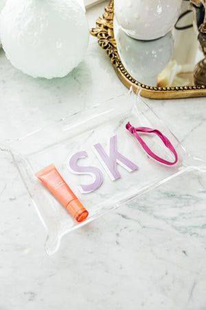 An acrylic tray with a purple and white monogram sits on a counter holding a hair tie and lip gloss