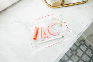 A large acrylic tray is customized with a pink and melon monogram and used to hold some jewelry and makeup products.