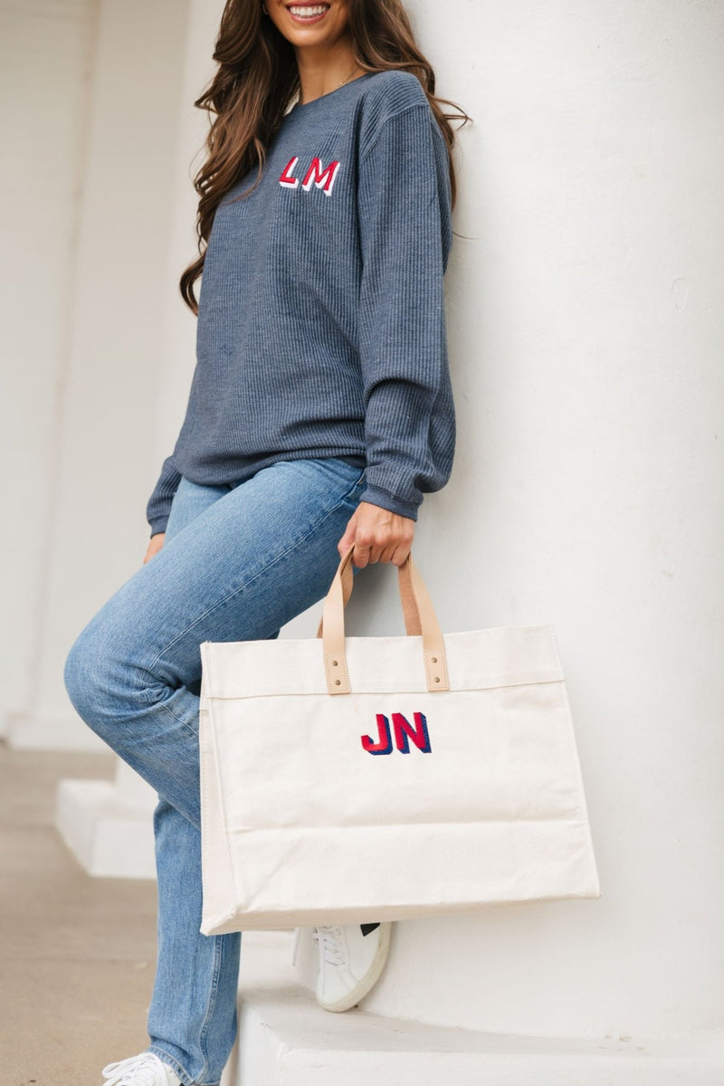 A brunette holds a monogrammed weekender while wearing a corded, monogrammed sweatshirts